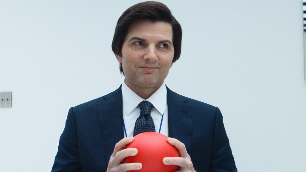 Adam Scott wearing a suit and holding a red ball, smiling, in Severance season 2.