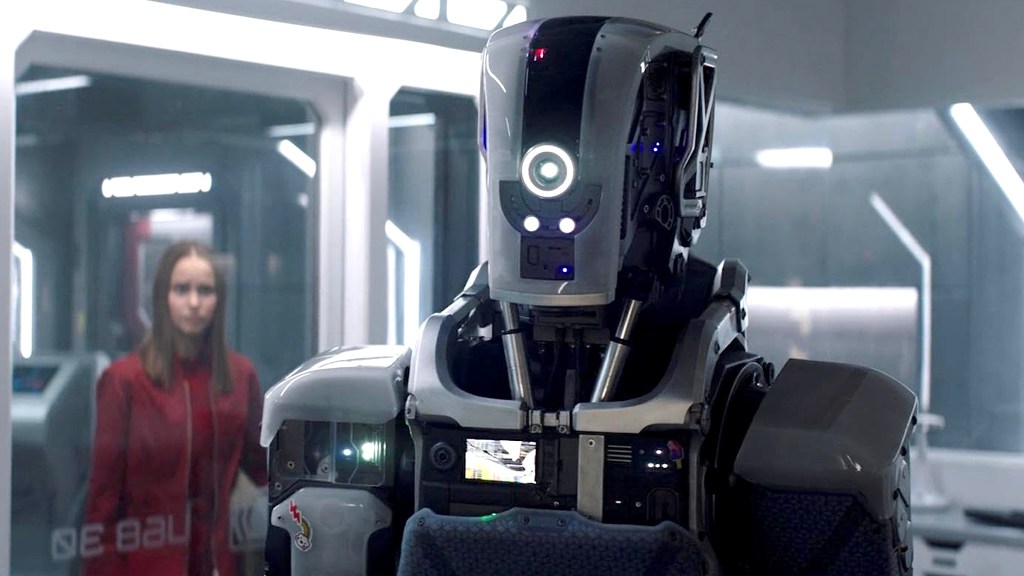 Un robot plateado con una chica vestida de rojo al fondo.