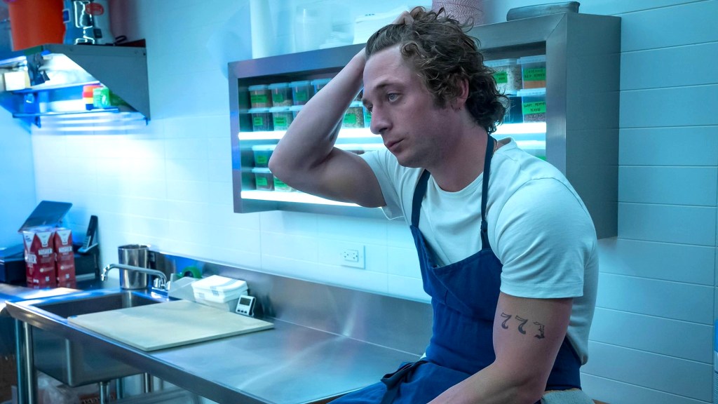 Jeremy Allen White as Carmen in The Bear wearing white t-shirt and blue apron, sitting on counter looking exhausted with his hand on his head