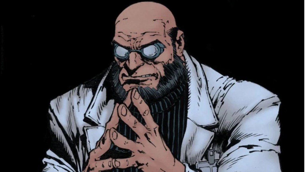 Hugo Strange sitting down with his hands steepled in front of him