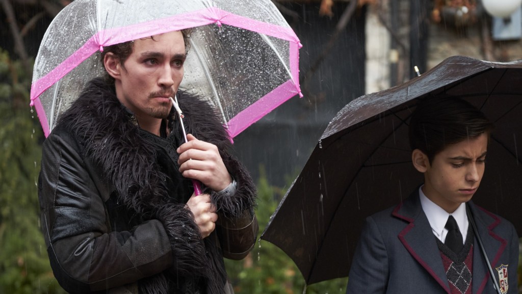 Robert Sheehan as Klaus in The Umbrella Academy holding a clear and pink umbrella